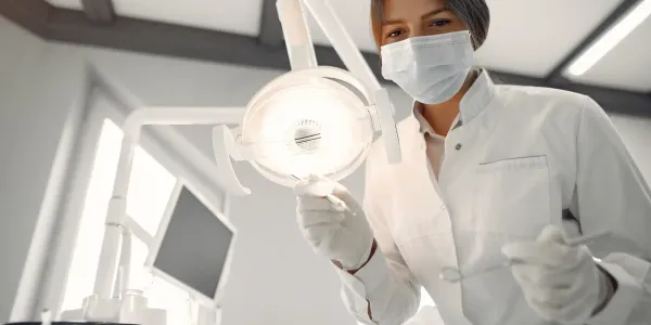 Profissional de saúde com máscara facial e luvas em ambiente clínico, segurando uma luz cirúrgica, representando área de saúde ou procedimentos médicos.